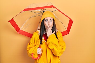Young hispanic woman wearing yellow raincoat holding umbrella covering mouth with hand, shocked and afraid for mistake. surprised expression