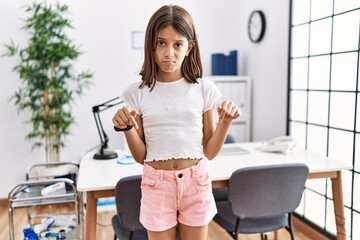 Young hispanic girl standing at pediatrician clinic pointing down looking sad and upset, indicating...
