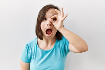 Young down syndrome woman standing over isolated background doing ok gesture shocked with surprised face, eye looking through fingers. unbelieving expression.