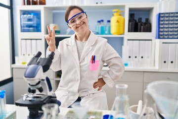 Fototapeta premium Hispanic girl with down syndrome working at scientist laboratory smiling looking to the camera showing fingers doing victory sign. number two.