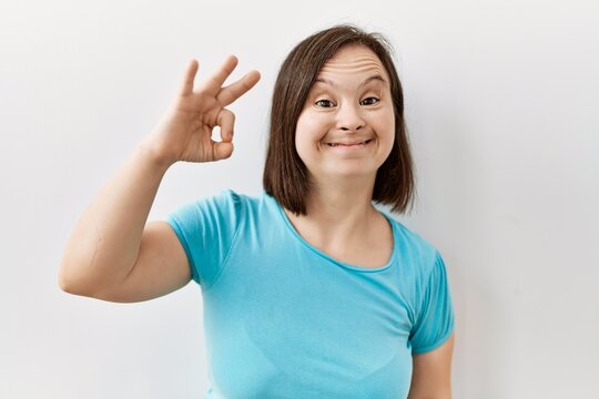 Young down syndrome woman standing over isolated background smiling positive doing ok sign with hand and fingers. successful expression.