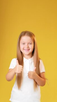 Satisfied child. Like gesture. Good idea. Approval sign. Vertical portrait of happy enthusiastic cheerful girl in white t-shirt showing thumbs up smiling isolated on yellow copy space background.