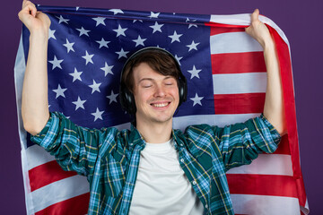 Happy man in a shirt holds an American flag and listens to music with headphones on a purple background. The celebration of Independence Day on July 4.