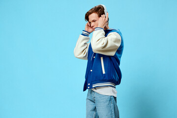 stylish guy listens to music in his white large headphones, holding them with his hands, standing on a light blue background in a stylish bomber jacket and jeans. Horizontal photo with space for text