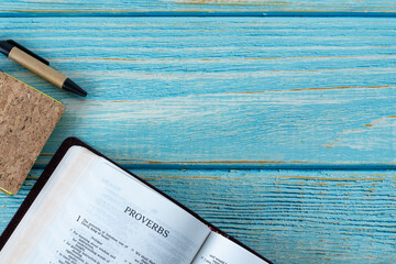 Proverbs open Holy Bible Book on a blue wooden background with a notebook and pen. Top table view....