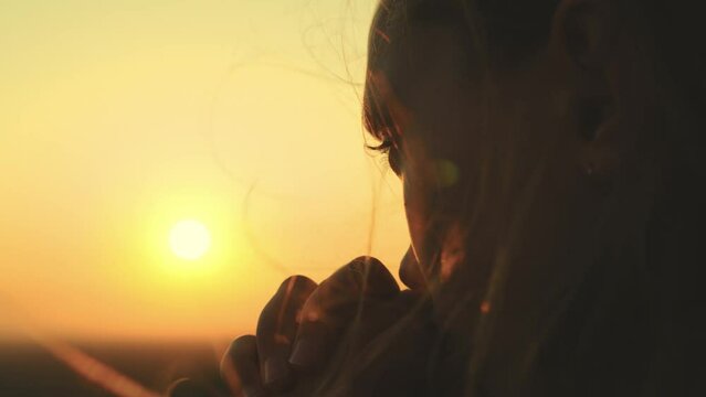 girl prays looking sunset, long hair flying away in glare sunlight rays strong wind, looking at dawn, lonely hike of brave girl, looking into sky with her eyes, believing good. close-up hands face.