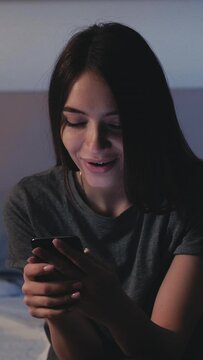 Digital life. Addicted woman. Mobile chatting. Sleepless smiling lady sitting bed scrolling smartphone feeling exhausted in light shadow room interior. Vertical video.