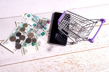 Still life: there is a grocery cart on a cell phone on the table and one thousand Russian ruble bills and coins scattered, the concept of shopping on the Internet