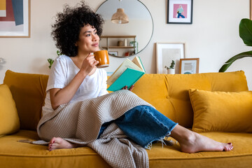 Pensive relaxed African american woman reading a book at home, drinking coffee sitting on the couch. Copy space. - obrazy, fototapety, plakaty