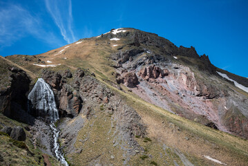 Summer mountain landscape with a waterfall and a stream. Picturesque mountain landscape of high mountains under a bright blue sky. Stunning mountain views.