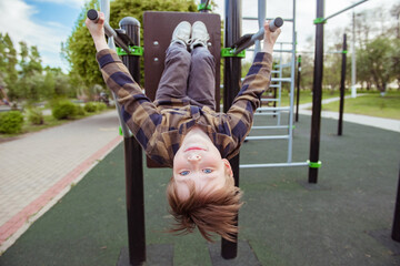 Outdoor portrait of attractive boy in the city playground. Happy kid hangs upside down on gym bars....