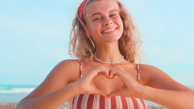 Close up portrait young happy Caucasian woman making shape heart gesture with hands and looking at you smiling, dressed in summer casual clothes stands on beach by sea. Volunteer, donation concept