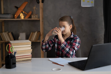 schoolgirl drinking water from a glass doing homework using a laptop, homeschooling, drinking...