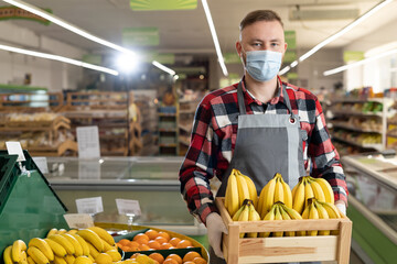 Charming salesman working in fruit section of supermarket. Supermarket clerk wearing a protective mask bringing a box of bananas