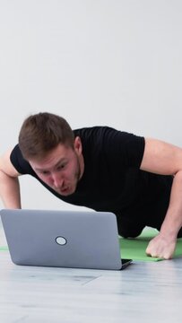 Hard workout. Exhausted man. Weak hands. Online training. Home gym. Tired guy doing push ups workout on fists looking laptop falling down yoga mat light room interior vertical.