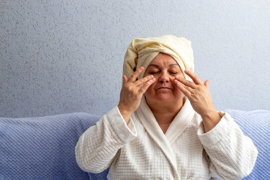 Elderly woman with towel on her head, does facial massage with two hands. Patches are pasted on the face. Selective focus. A picture for articles about age-related facial care.