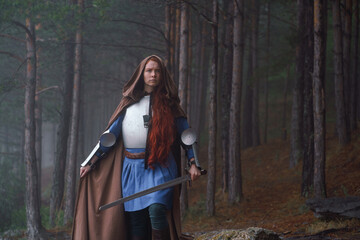 Fototapeta premium Red-haired girl in armor and raincoat in forest. Historical concept