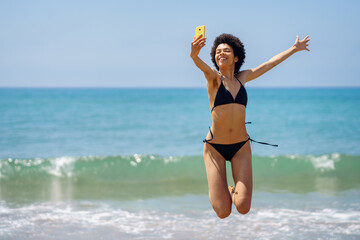 Happy black woman taking selfie while jumping on coast