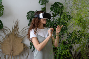 Home gardening, virtual technology, metaverse concept. Hispanic young woman touching asparagus houseplant in immersive space VR. Italian female using VR glasses in greenhouse. Augmented reality. 