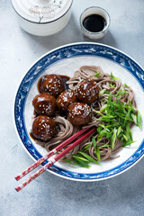 Plate of pork meatballs in teriyaki sauce with soba noodles, studio shot on a light-blue stone background, elevated view