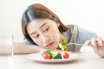 Obraz na płótnie Canvas Anorexia, unhappy beautiful asian young woman, girl on dieting, holding fork at broccoli in salad plate, dislike or tired with eat fresh vegetables. Nutrition of good healthy, bored with food.