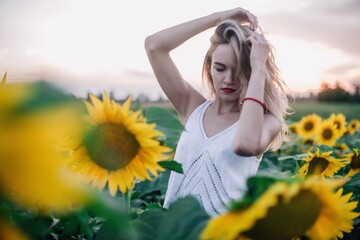 Fototapeta na wymiar A young, slender girl with loose hair in a T-shirt stands in a field of sunflowers at sunset