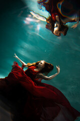 Photo underwater, a young beautiful woman in red with red hair reaches for the surface of the...