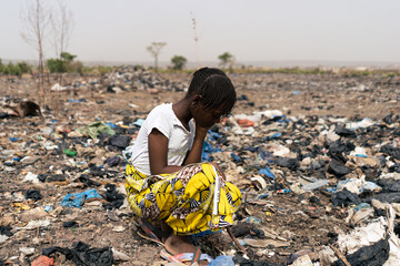 Exhausted little African girl kneeling in a garbage dump where she is forced to search for reusable...