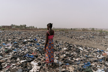 Lonely little African girl standing on a landfill in the midst of all types of garbage; environmental pollution concept