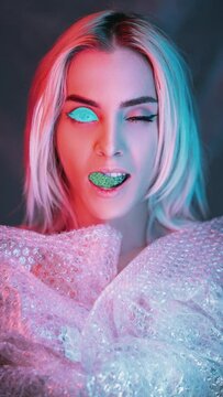NFT art. Digital beauty. Crypto portrait. Pink blue neon light cyborg girl face with flicker glitch noise winking eye open mouth in plastic bubble wrap isolated on gray vertical.
