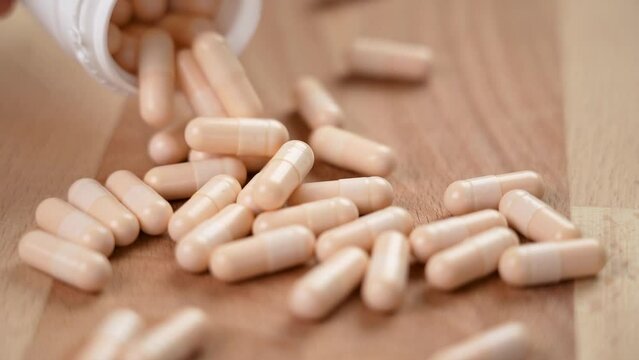 Vitamin B capsules fall from a white plastic container on a wooden eco surface in slow motion close up