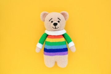 Crochet amigurumi handmade stuffed soft teddy bear toy in rainbow sweater on yellow background....