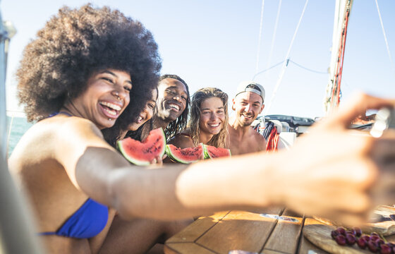 Cheerful multiracial people eating watermelon while girl with curly hair taking a selfie - Young friends having fun on boat during their holiday