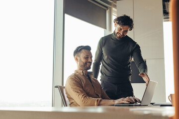 Obraz premium Positive male colleagues discussing common project, using laptop, brainstorming for ideas at modern office, empty space