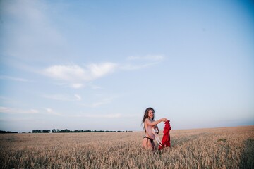 A young, slender girl with loose hair in a field of wheat hides behind a hat and tosses a red dress.