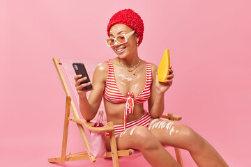 Happy woman applies sun cream gets suntan while sitting on deck chair holds bottle of sunscreen lotion for skin care uses smartphone for checking newsfeed dressed in swimwear isolated over pink wall