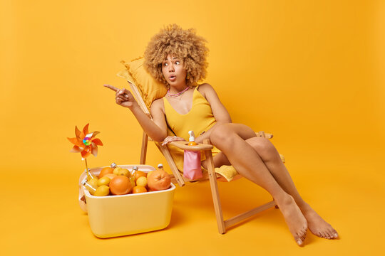 Shocked curly haired young woman dressed in swimsuit points left with index finger poses on comfortable deck chair near portable freezer with citrus fruits isolated over vivid yellow background