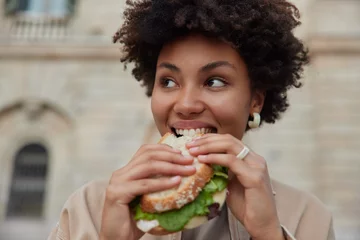 Tuinposter Pretty curly haired woman bites delicious sandwich poses outdoors at street looks away dressed casually has quick snack while walking outside being hungry. People lifestyle and fast food concept © Wayhome Studio
