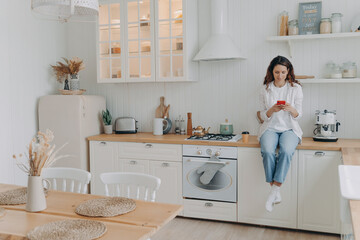 Fototapeta na wymiar Happy young woman has message on phone. Girl sitting on worktop at kitchen at her apartment
