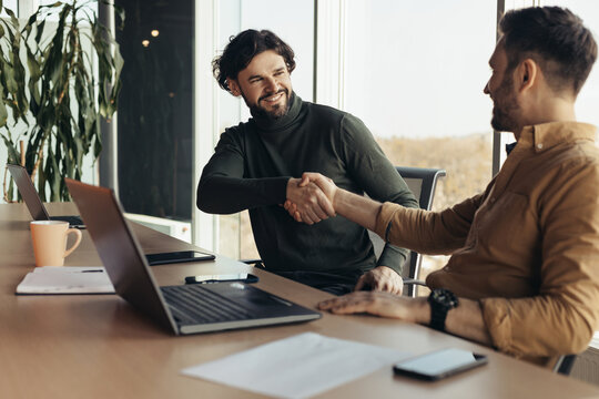 Happy business partners shaking hands in agreement, making deal, sitting at desk with laptop at modern office