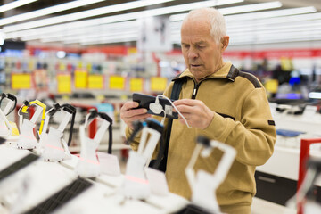 elderly grayhaired man pensioner examining counter with electronic gadgets and tablets in showroom...
