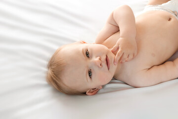 Happy naked baby in diaper lying on his back on the white sheet and touching his face. Baby girl with red hair looking at camera. Light background. Little child. Serious emotion. Advertising Mockup