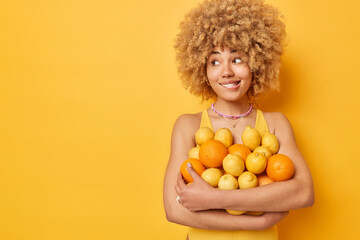 Fototapeta na wymiar Beautiful curly haired woman keeps to fruit diet carries fresh oranges and lemons bites lips looks away dressed in swimwear isolated over vivid yellow background copy space for your promotion