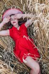 A young, slender, sexy girl in a bright red dress and hat lies in a field of wheat