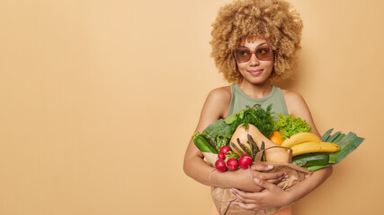 Horizontal shot of thoughtful curly haired woman holds paper bag full of fresh fruits and vegetables from supermarket eats healthy food full of vitamins wears sunglasses isolated over beige background