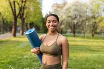 Fototapeta Happy young black woman in sports clothes holding fitness mat, smiling at camera, ready for yoga practice at urban park obraz