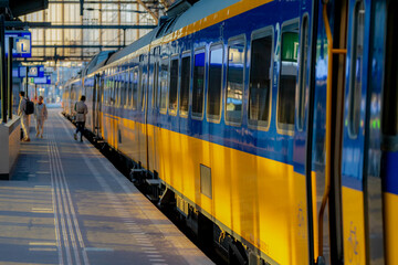 Naklejka na ściany i meble Selective focus of the train with blurred people traveling as background, Yellow blue bogie and passenger getting in and out the train in public station, Netherlands.