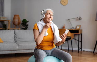 Happy senior woman with wireless headset sitting on fitball, listening to music on phone, resting...