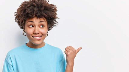 Curious pretty young woman with curly hair bites lips points thumb away on empty space dressed in casual blue t shirt shows advertisement or promo isolated over white background. Follow this way