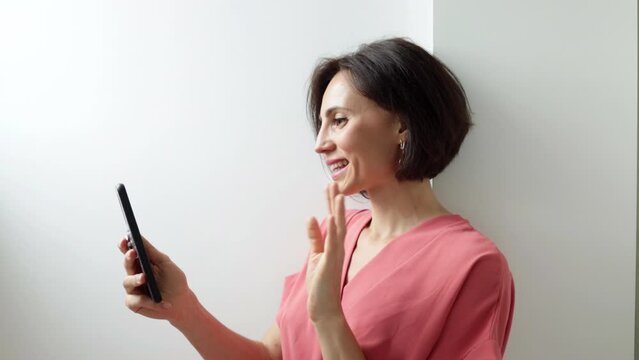 Smiling millennial female holding smartphone talking by video call wave hand to camera. Positive young mixed race woman contact call friend by video link chat using device webcam
